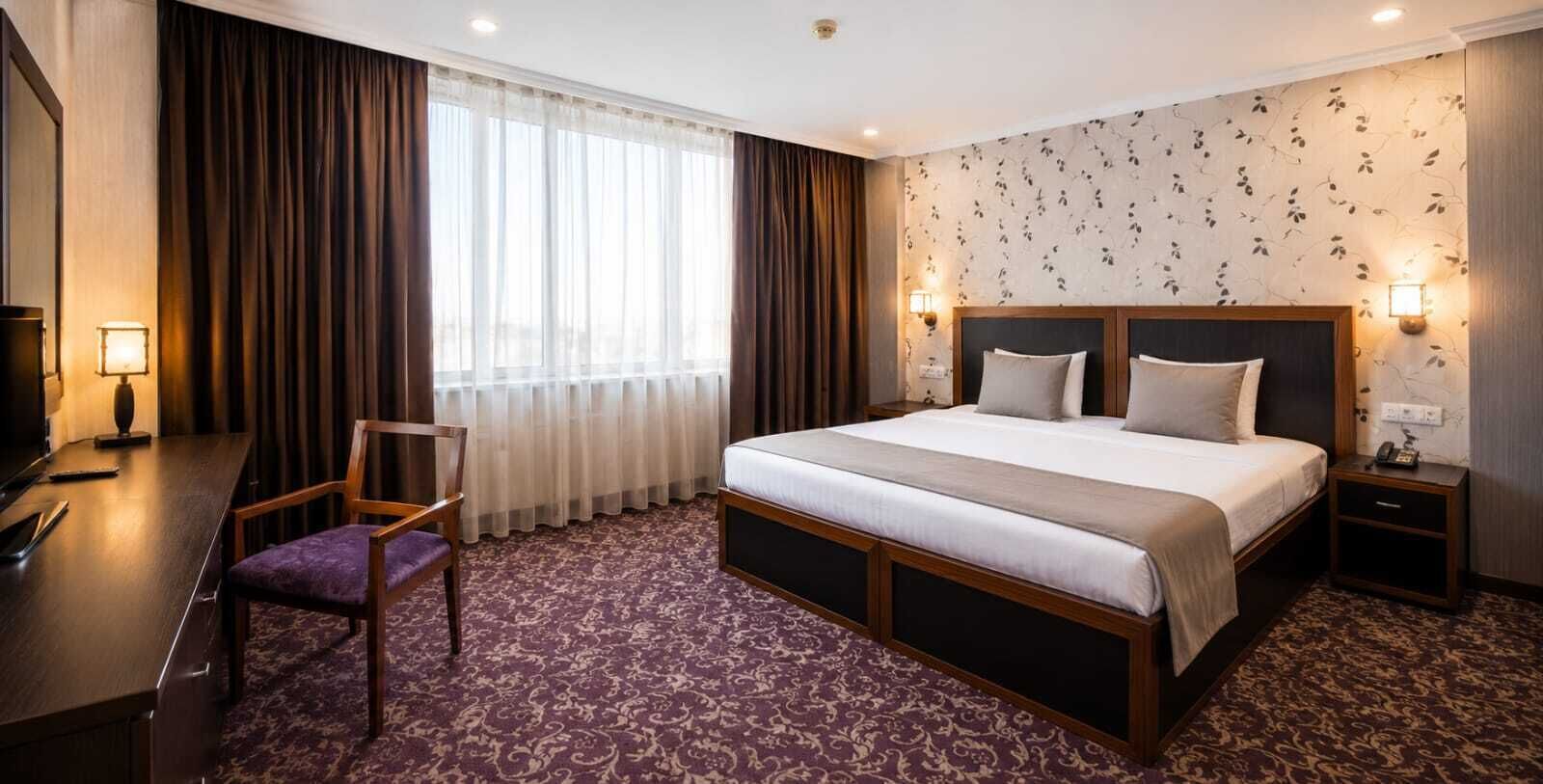 A hotel room with a large bed, a desk with a chair, a TV, and a nightstand with a lamp.