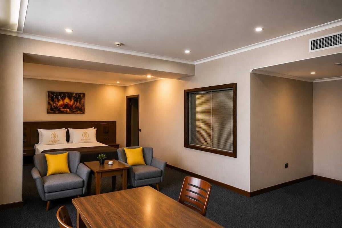 A hotel room with a bed, two armchairs, a wooden table, a painting on the wall, and a window with blinds.