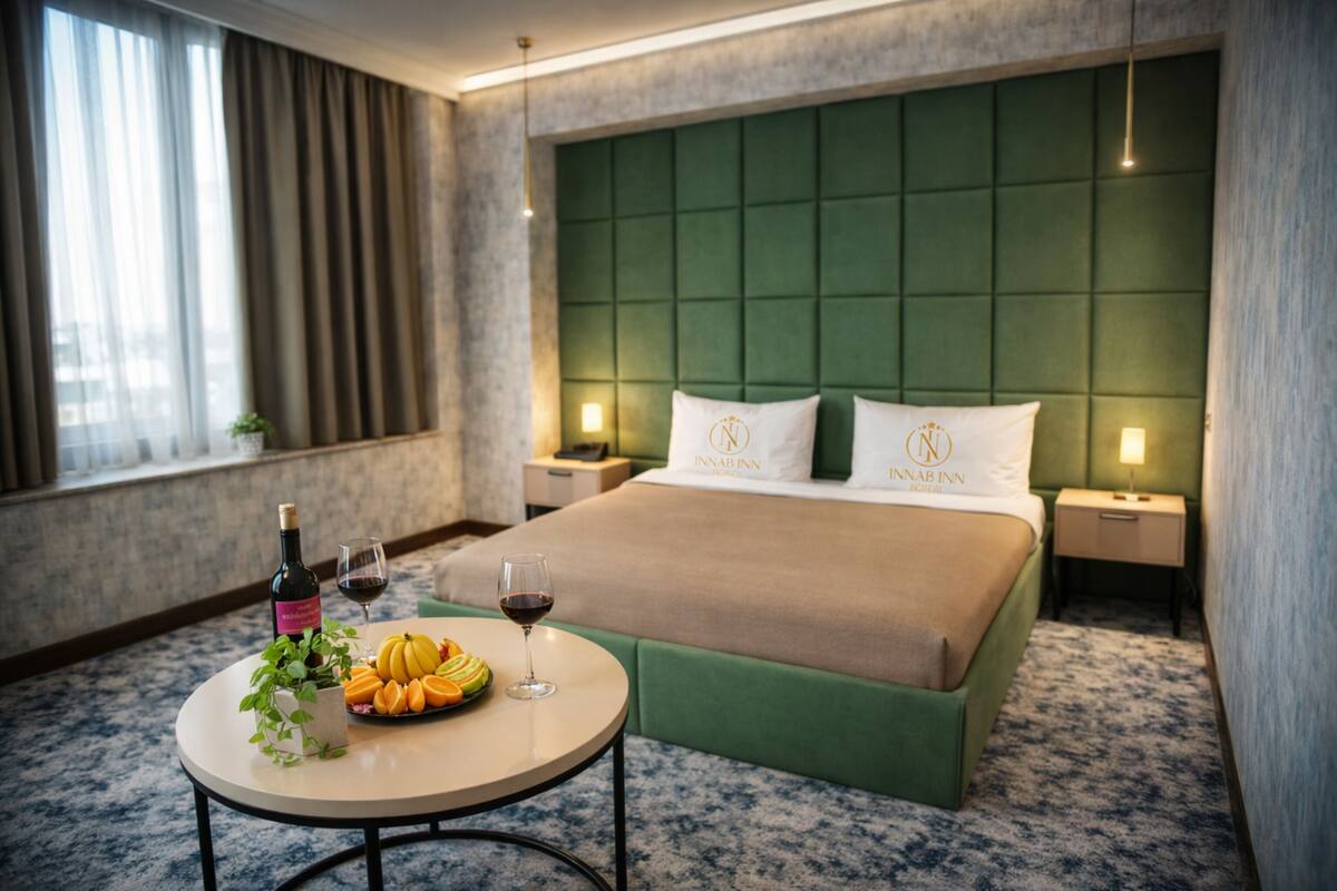 A hotel room with a green upholstered headboard, a bed with two pillows, a bedside table with a bottle of wine and a glass, and a fruit basket on the floor.
