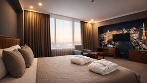 Habitación de hotel con una cama grande, dos almohadas, un escritorio, una silla, un televisor y un cuadro de un paisaje urbano.