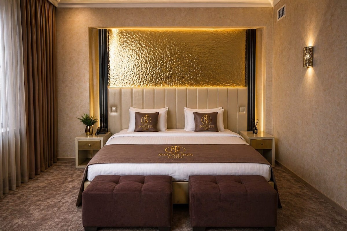 A hotel room with a large bed, two bedside tables, a headboard with a textured gold panel, and a wall-mounted light fixture.