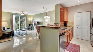 Una cocina moderna con una isla central, un comedor con sofá y mesa de centro, y vistas al exterior a través de grandes ventanales.
