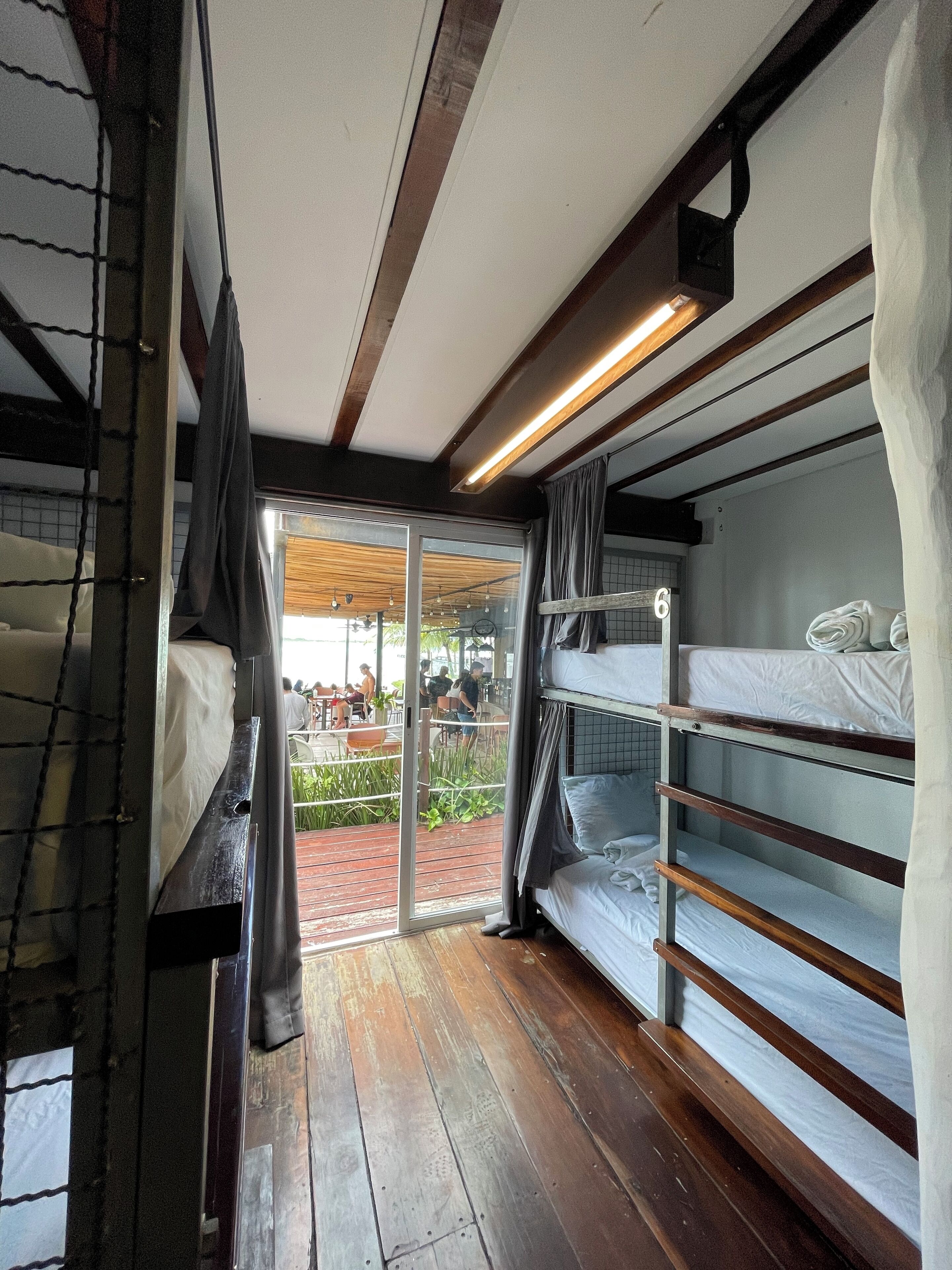 A room with bunk beds, a wooden floor, and a sliding glass door leading to an outdoor area with tables and chairs.