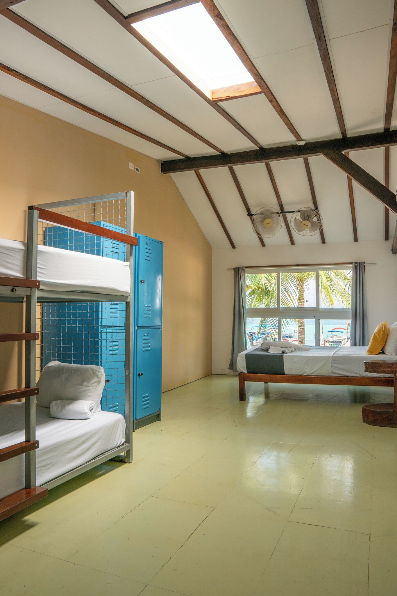 A dormitory room with bunk beds, a window with a view of palm trees, and a ceiling fan.