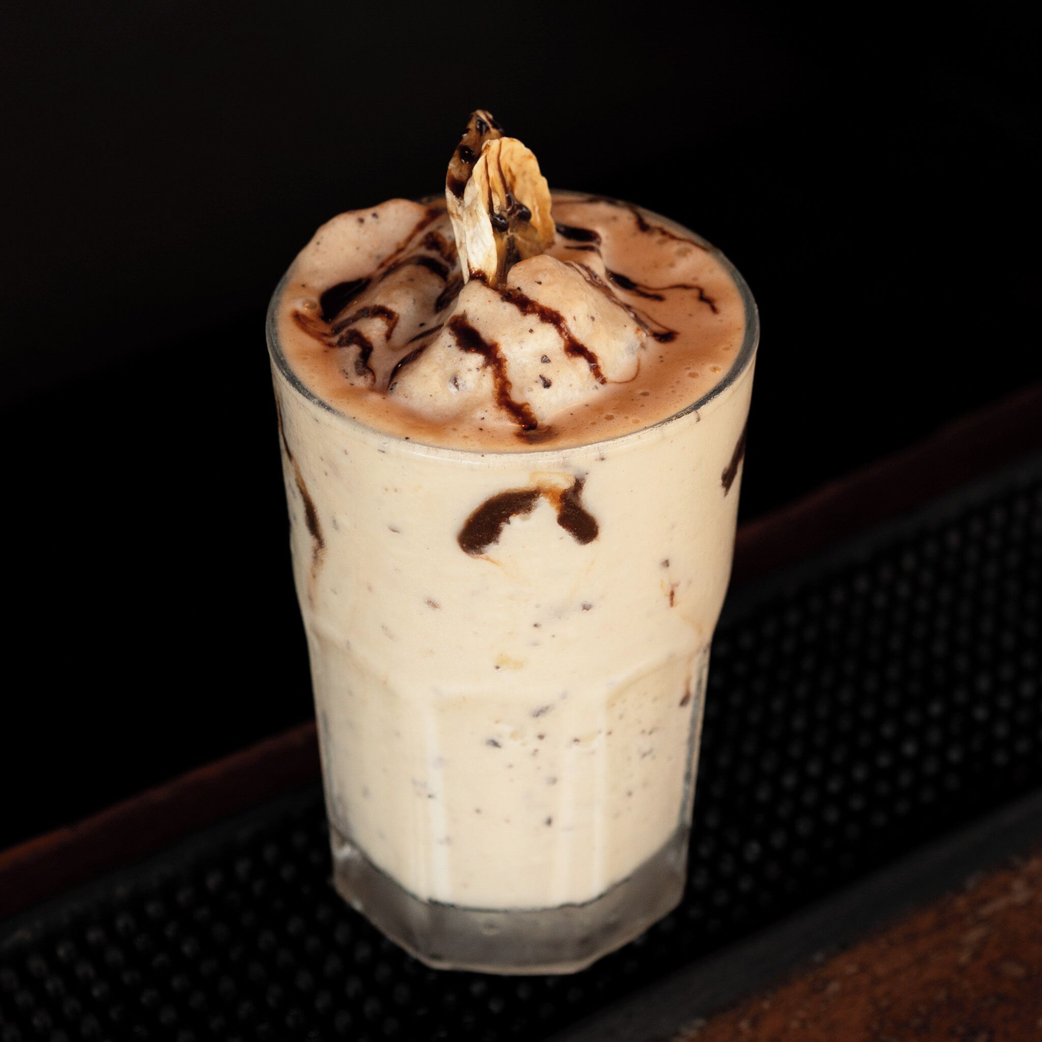 A glass of layered beverage with chocolate syrup on a dark surface.