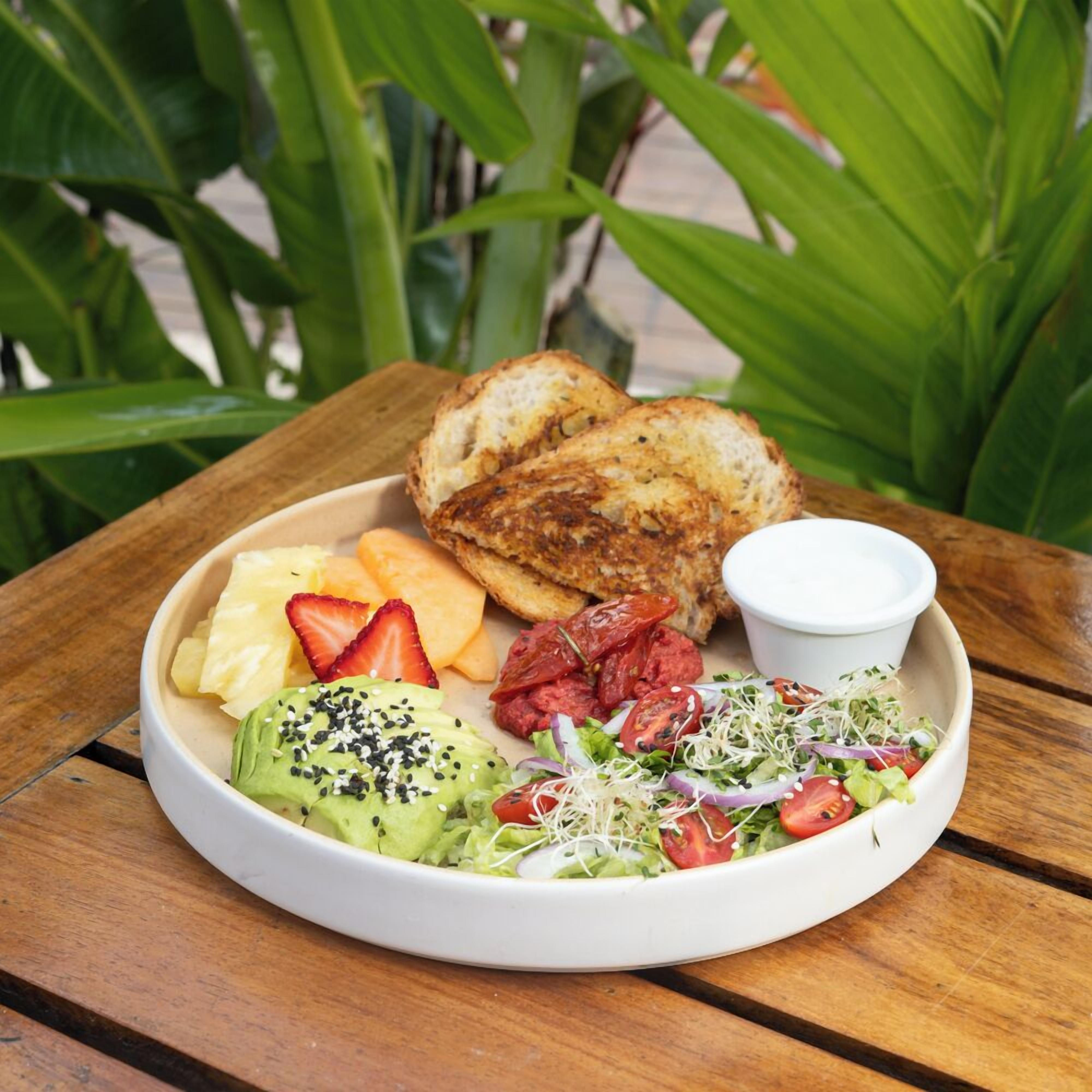 Un repas composé de pain grillé, d’avocat, de fraises, de fromage et d’une salade agrémentée de jeunes pousses et de tomates, servi sur une table en bois.