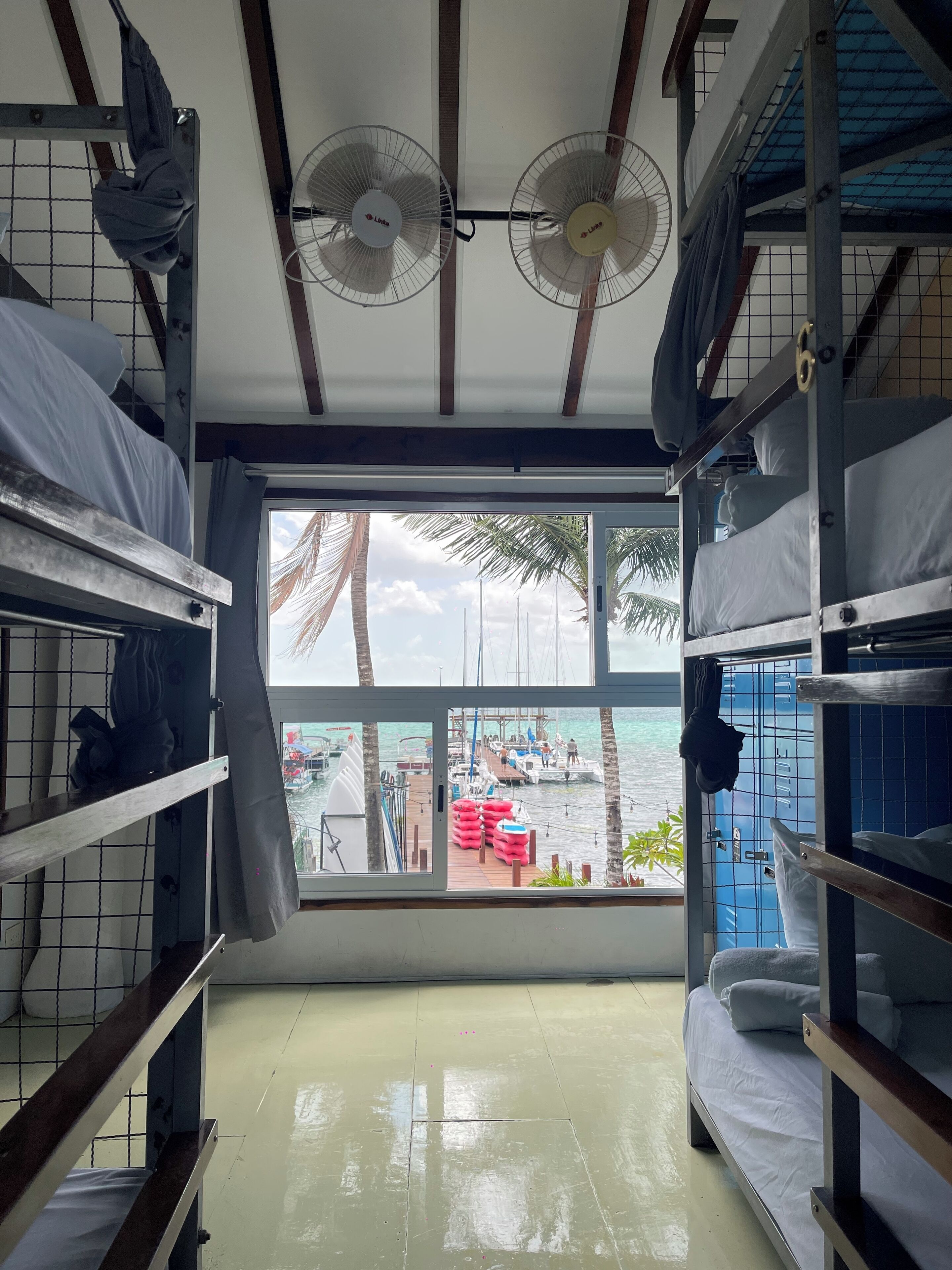 Una habitación con literas, ventana con vista a barcos y palmeras, y dos ventiladores de techo.