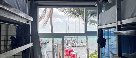 Una habitación con literas, ventana con vista a barcos y palmeras, y dos ventiladores de techo.