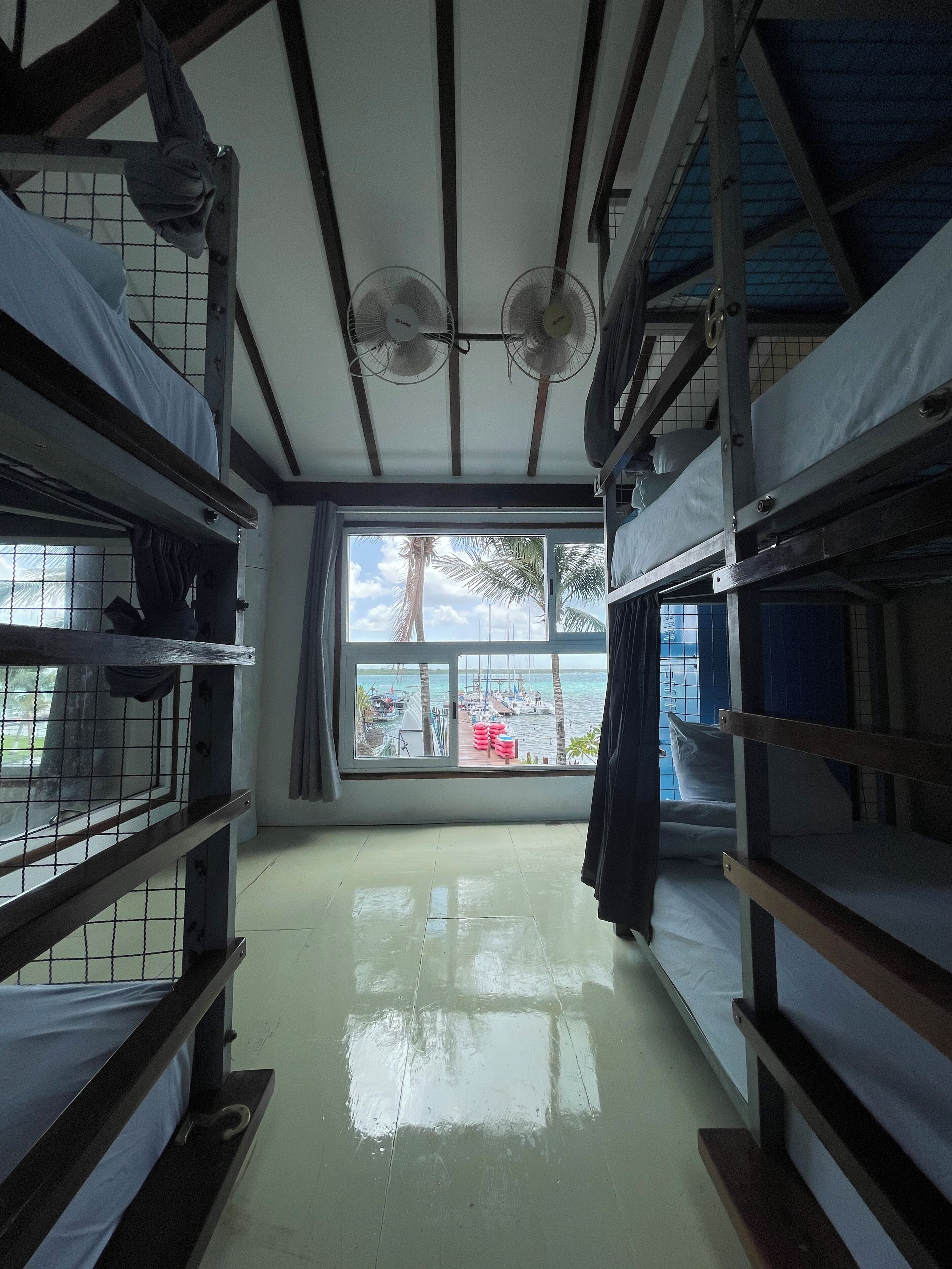 A dormitory room with bunk beds, a large window with a view of boats and palm trees, and ceiling fans.