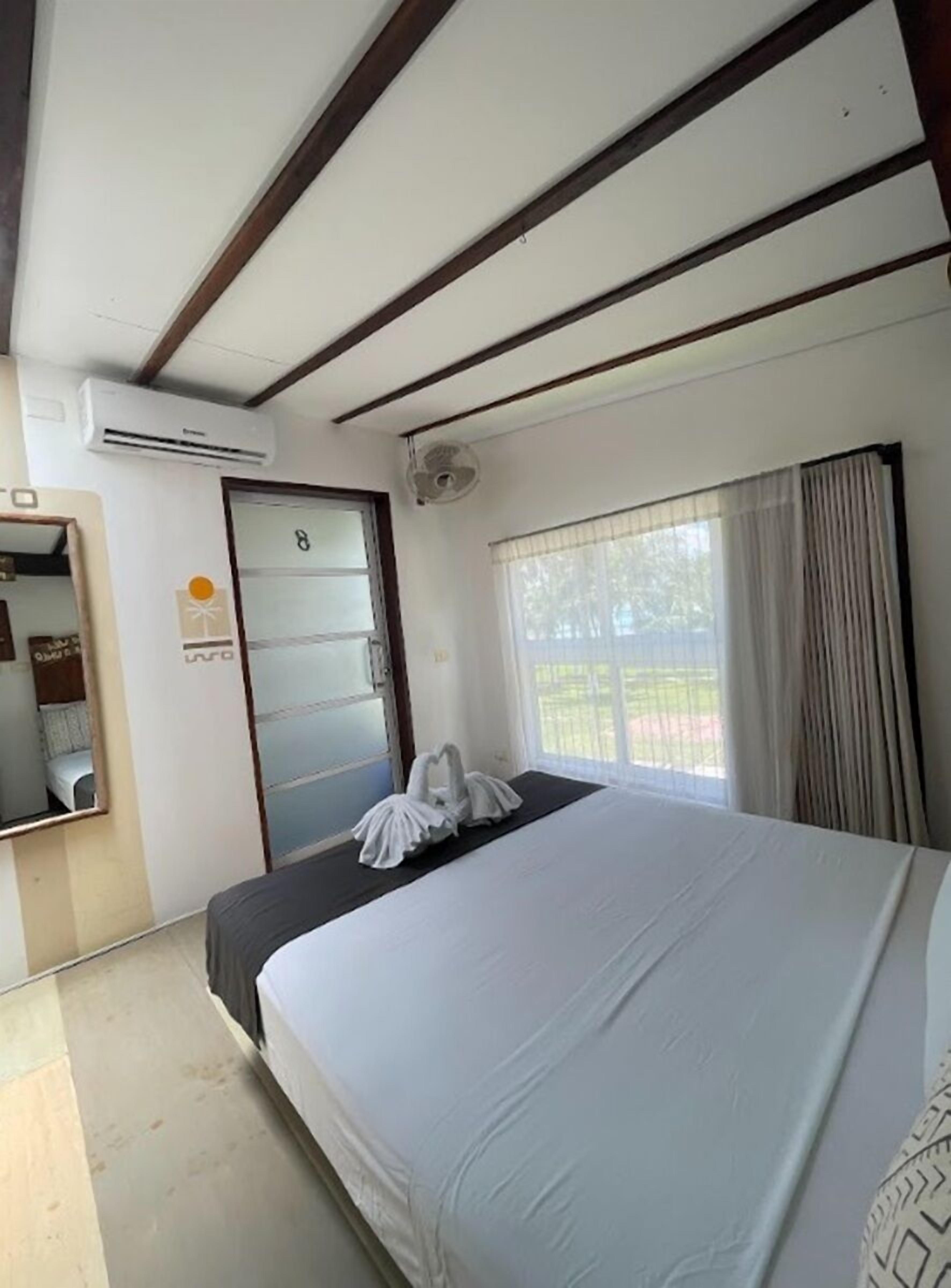 A hotel room with a bed, a window with curtains, an air conditioning unit, and a door with a number.