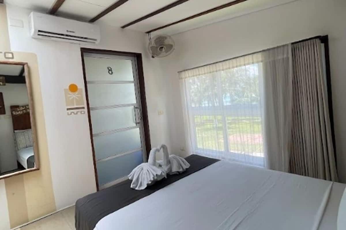 A hotel room with a bed, a window with curtains, an air conditioning unit, and a door with a number.