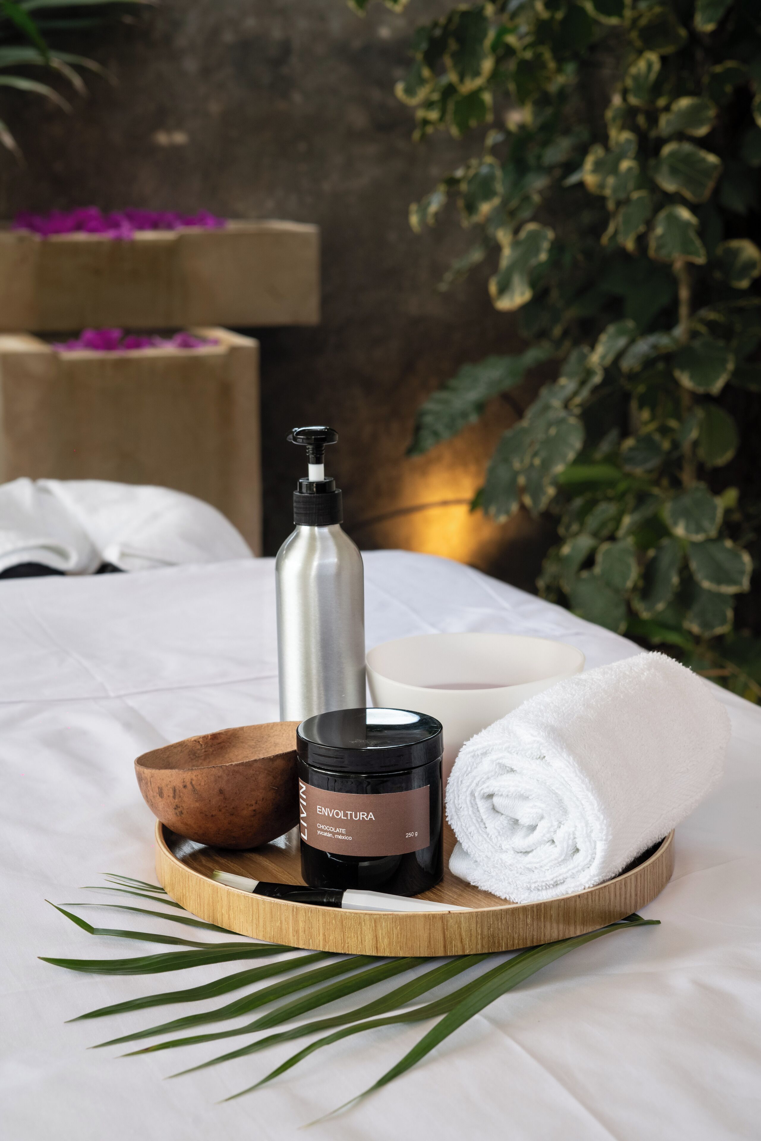 A wooden tray with a black bottle, a white towel, a wooden bowl, and a white candle on a bed with a white sheet.