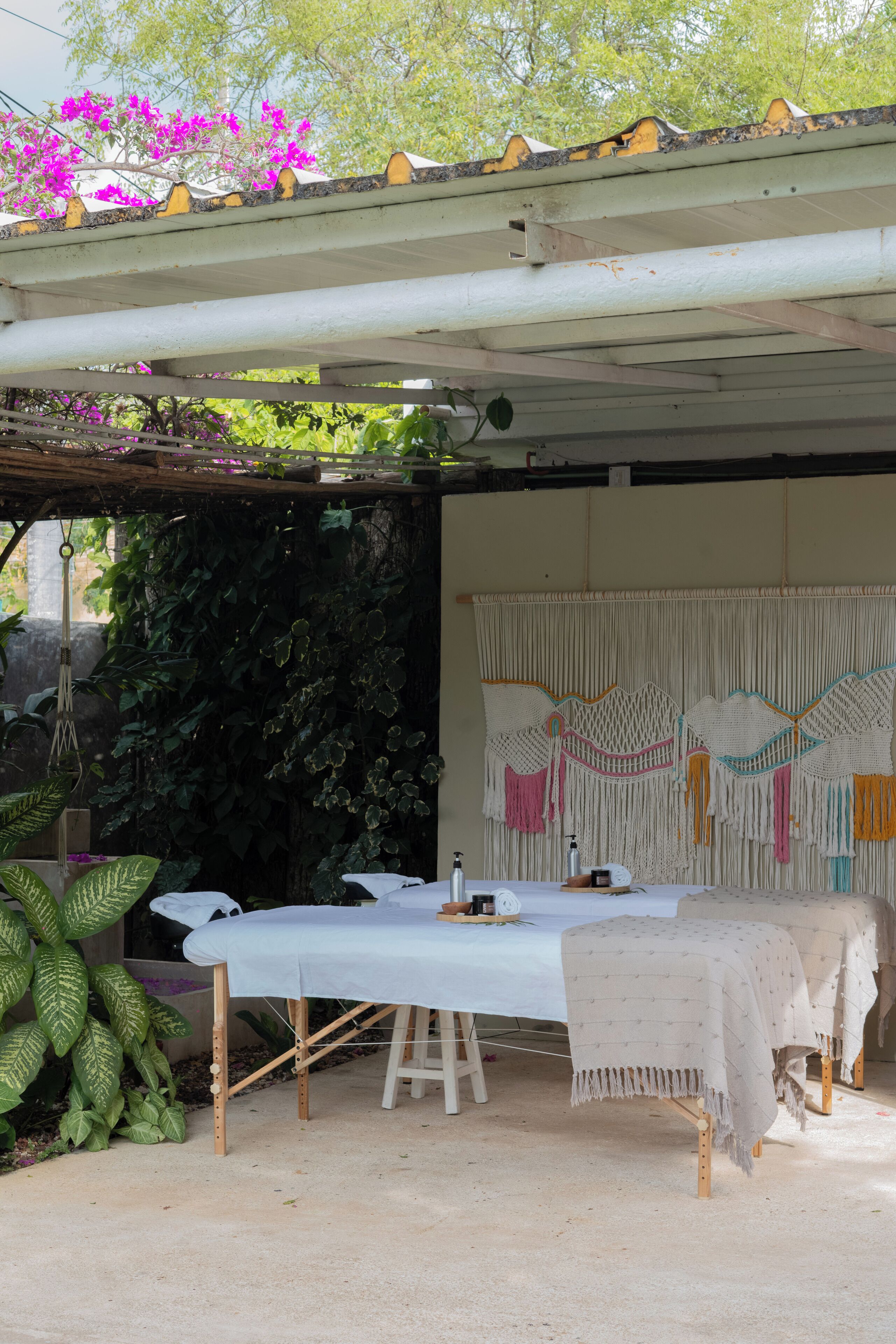 A covered outdoor seating area with a table and benches, surrounded by greenery and a decorative wall.