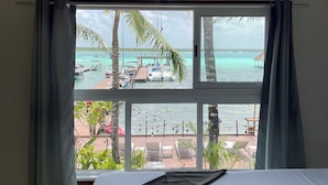Et hotelværelse med seng, to loftsvifter, et stort vindue med udsigt til strand og både samt en balkon med stole og planter.