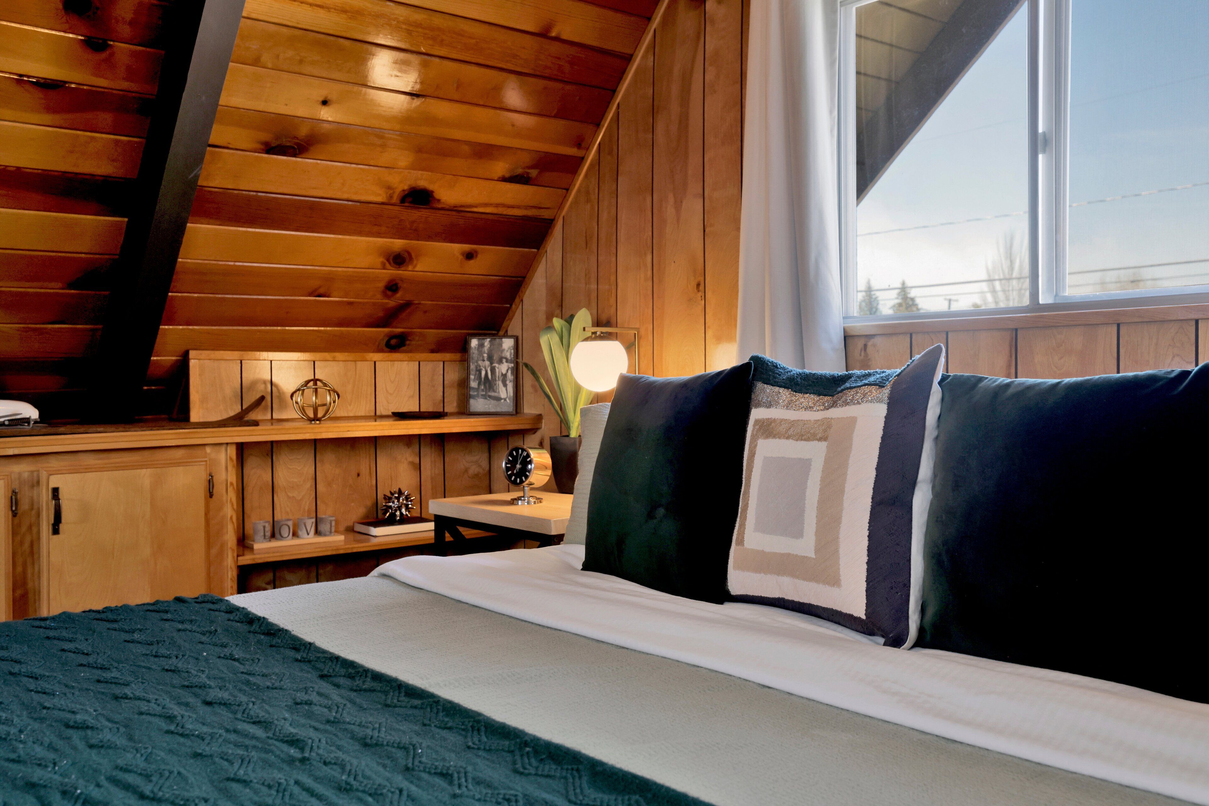 Interior de una cabaña de madera con una cama, una mesita de noche, una lámpara y una ventana.