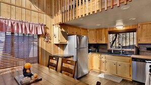 A wooden cabin-style kitchen with a refrigerator, wooden cabinets, a dining table, and a window with blinds. - Black Diamond Vista #653 (Big Bear Lake)