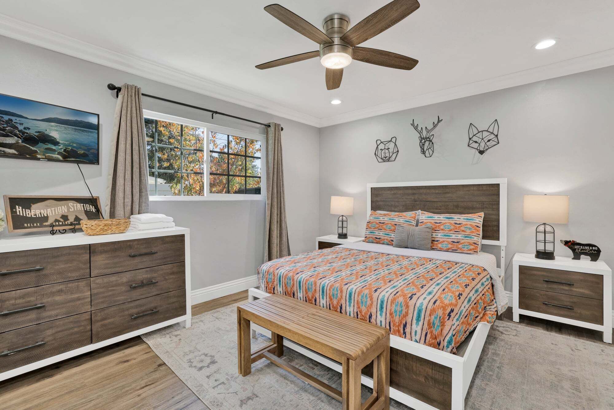 A bedroom with a bed, bedside tables, a bench, a ceiling fan, a window with curtains, and wall art.