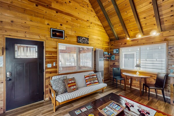 A cozy cabin interior with a wooden ceiling, a sofa with patterned cushions, a dining table with chairs, and a window with blinds. - Bristlecone Cottage #321 (Big Bear Lake)