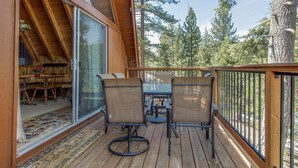 A wooden deck with a table and chairs, a glass door leading to an interior space, and a view of tall trees. - Castle Rock Cabin #2 (Big Bear Lake)
