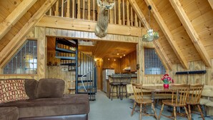 A cozy loft-style room with a wooden staircase, a mounted deer head, a dining area with wooden chairs, and a sofa with patterned cushions. - Castle Rock Cabin #2 (Big Bear Lake)