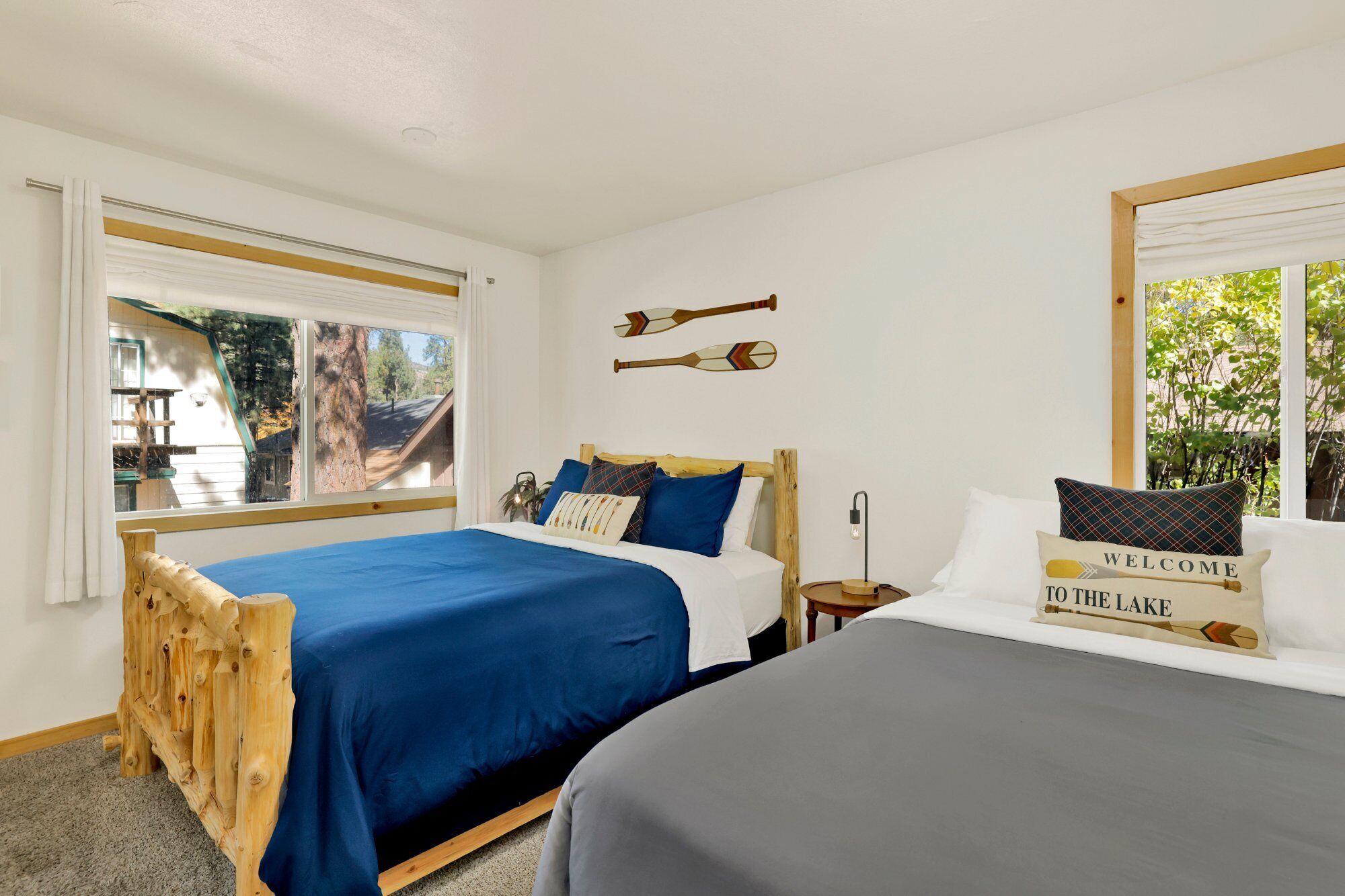 A bedroom with two beds, a wooden headboard, a nightstand, and a window with a view of trees and a building.