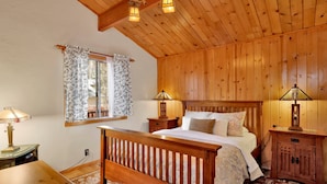 A wooden-paneled bedroom with a bed, two bedside tables with lamps, a nightstand, and a window with floral curtains. - Craftsman Hideaway #630 (Big Bear Lake)