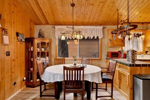 Un intérieur en bois avec une table de salle à manger ronde, des chaises, un buffet aux portes vitrées, une fenêtre ornée de rideaux en dentelle et un coin cuisine équipé d’un four à micro-ondes et d’un réfrigérateur.