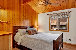 Une chambre à coucher dans une cabane en bois, avec un lit, une table de chevet, une fenêtre avec un rideau et un ventilateur de plafond.