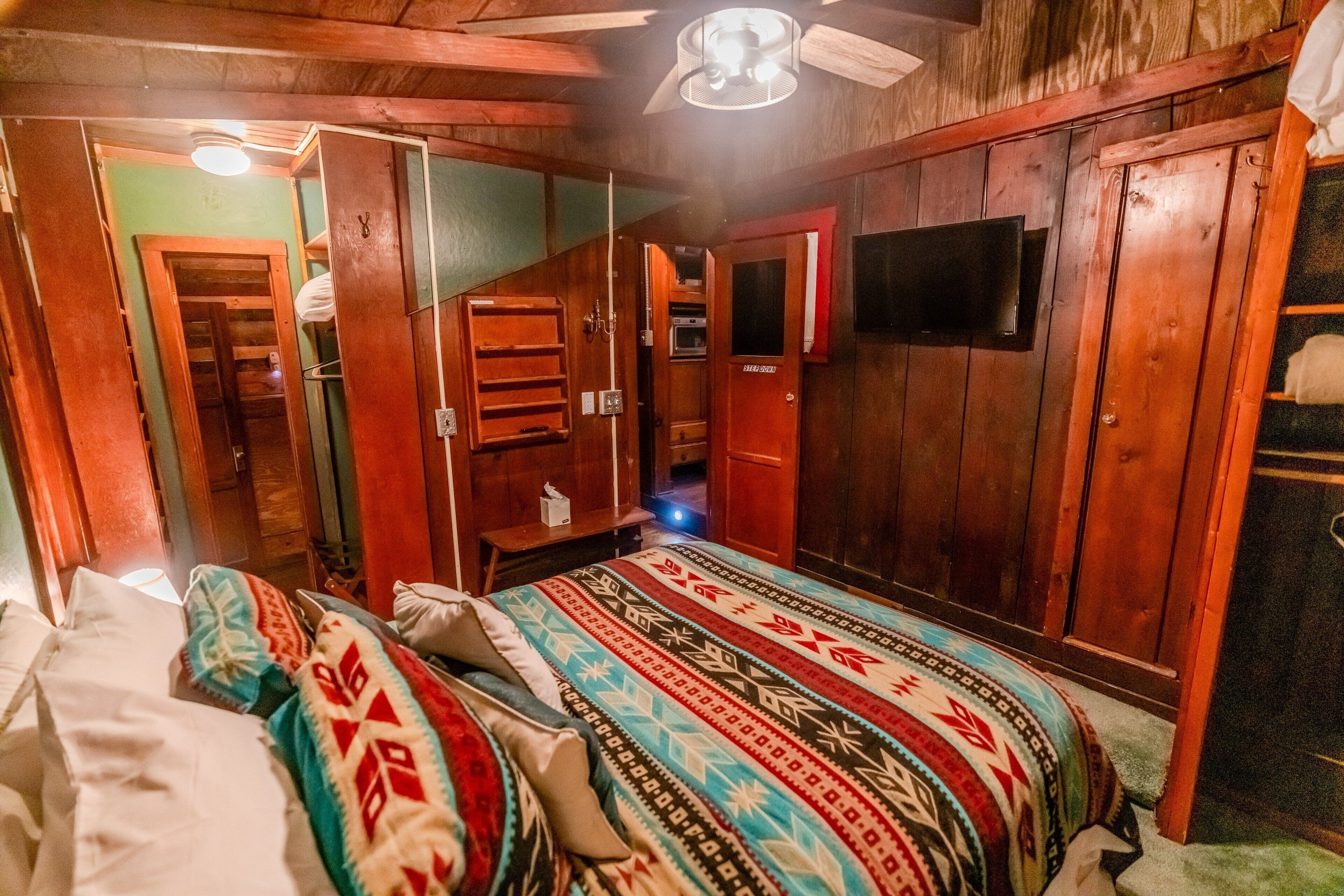 A room with wooden walls, a bed with patterned bedding, a television mounted on the wall, and a ceiling fan.