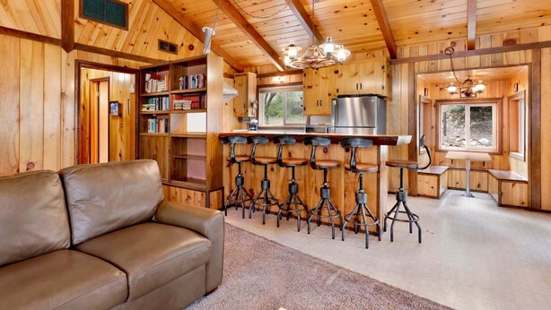 A wooden interior with a sofa, bar stools, a bookshelf, and a kitchen area. - Idle Ours Cabin #713 (Fawnskin)