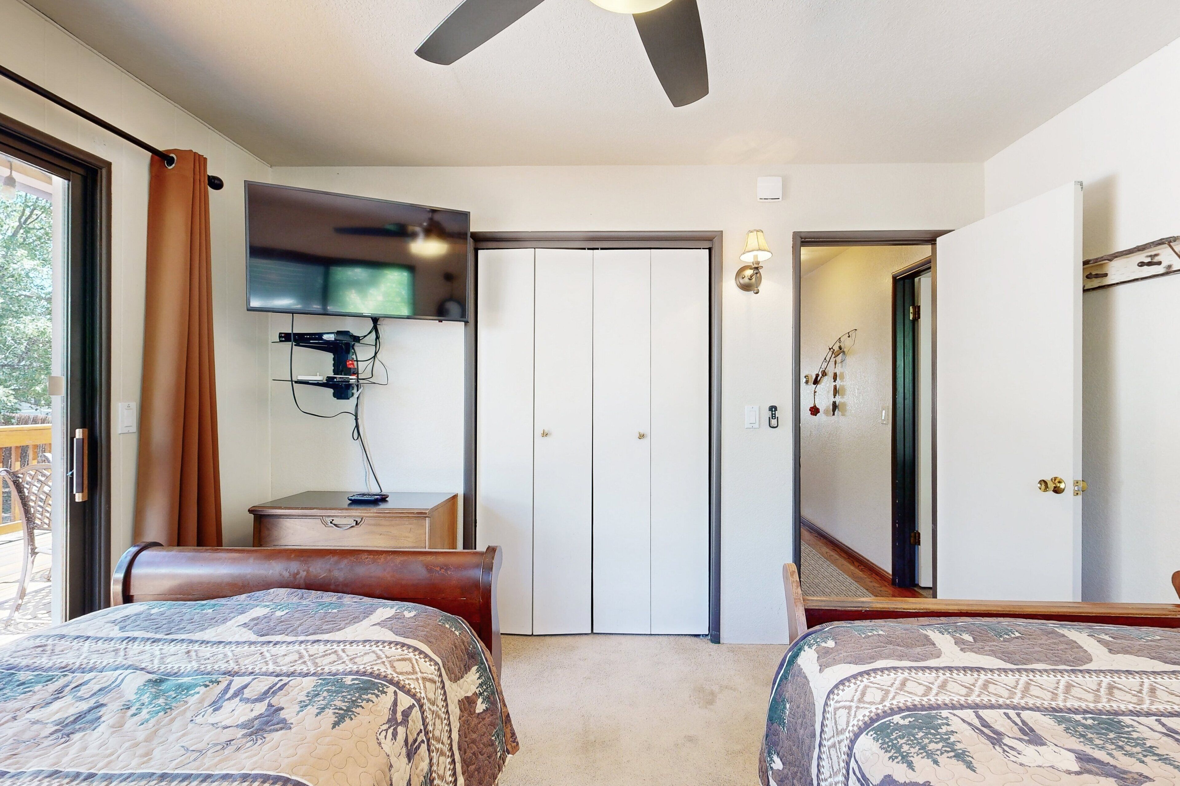 Ein Hotelzimmer mit zwei Betten, einem Deckenventilator, einem an der Wand montierten Fernseher, einer Tür zu einem Flur und einem kleinen Schreibtisch mit Lampe.