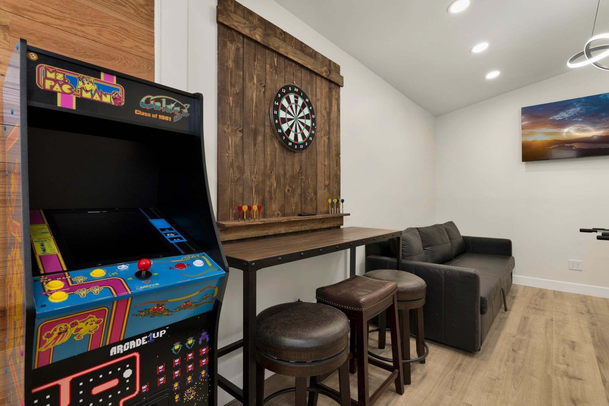 Una habitación con una máquina de videojuegos retro, un dardo, un sofá y un cuadro en la pared.