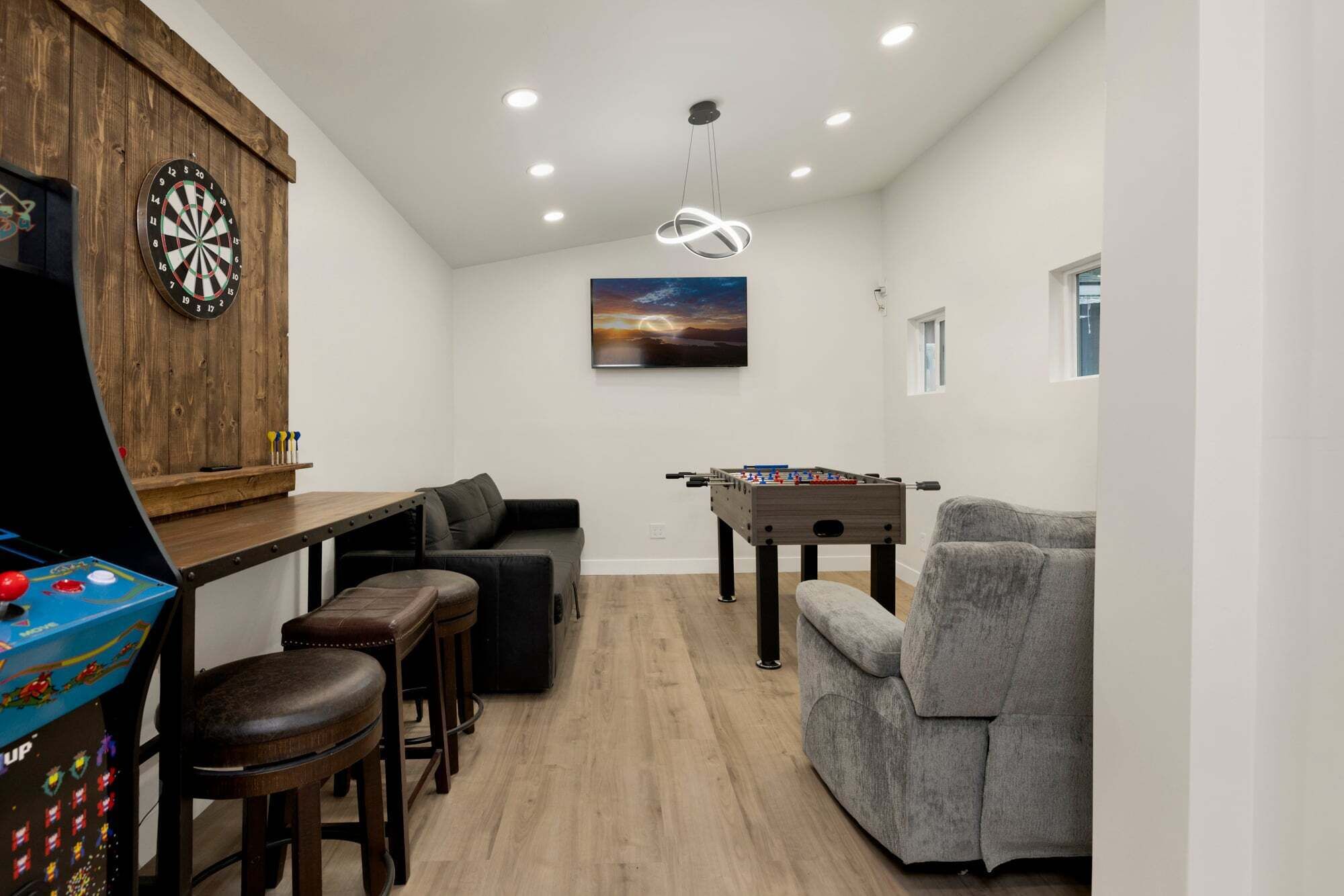 A room with a dartboard, a foosball table, a bar area with stools, and a grey armchair.