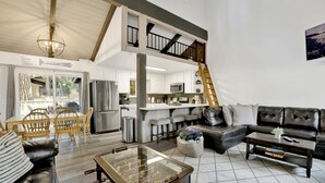A spacious living room with a high ceiling, a built-in kitchen, a dining area, and a cozy seating area with a fireplace. - Moose-in' Around (Big Bear Lake)