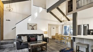 A spacious living area with a leather sofa, a coffee table, and a dining table with chairs. A kitchen area with a refrigerator and a coffee maker is visible in the background. - Moose-in' Around (Big Bear Lake)