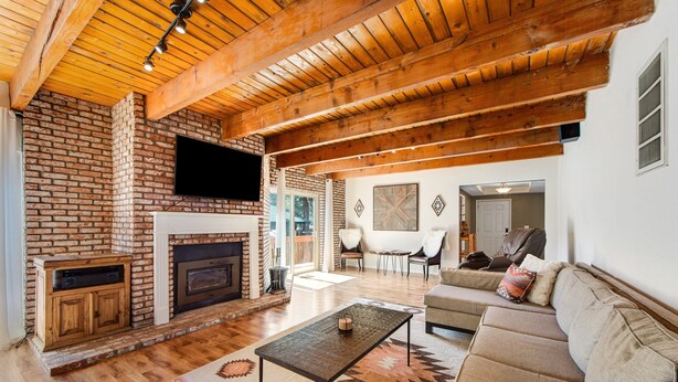 A spacious living room with a brick fireplace, wooden ceiling beams, and a large sofa. - Mt Doble Dream Home #504 (Big Bear City)