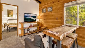 A cozy cabin interior with a wooden interior, a television on a wooden stand, a dining table, and a view of greenery outside. - Orchard Family Cabin #2560 (Big Bear Lake)
