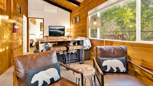 A cozy cabin interior with wooden walls, a large window, a television, and two chairs with bear-themed cushions. - Orchard Family Cabin #2560 (Big Bear Lake)