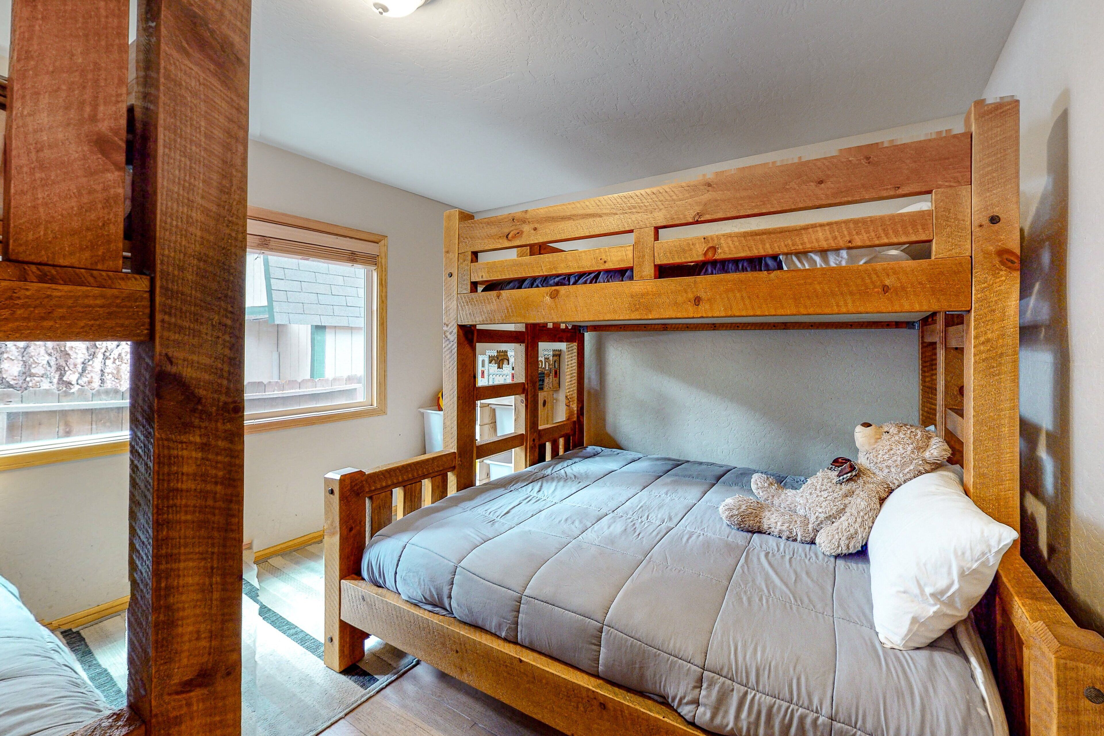 A bunk bed with a teddy bear on the top bunk, a window with blinds, and a wooden headboard.