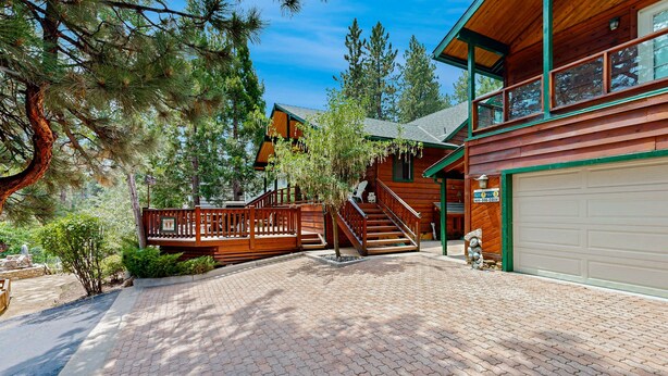 A two-story log cabin with a brick driveway, wooden deck, and a garage. - Rooster's Den #710 (Big Bear Lake)