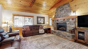 A cozy living room with a stone fireplace, wooden ceiling, and a large flat-screen TV.