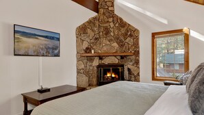 Une chambre avec une cheminée en pierre, un lit recouvert d’une courtepointe, une table de chevet et un tableau accroché au mur.