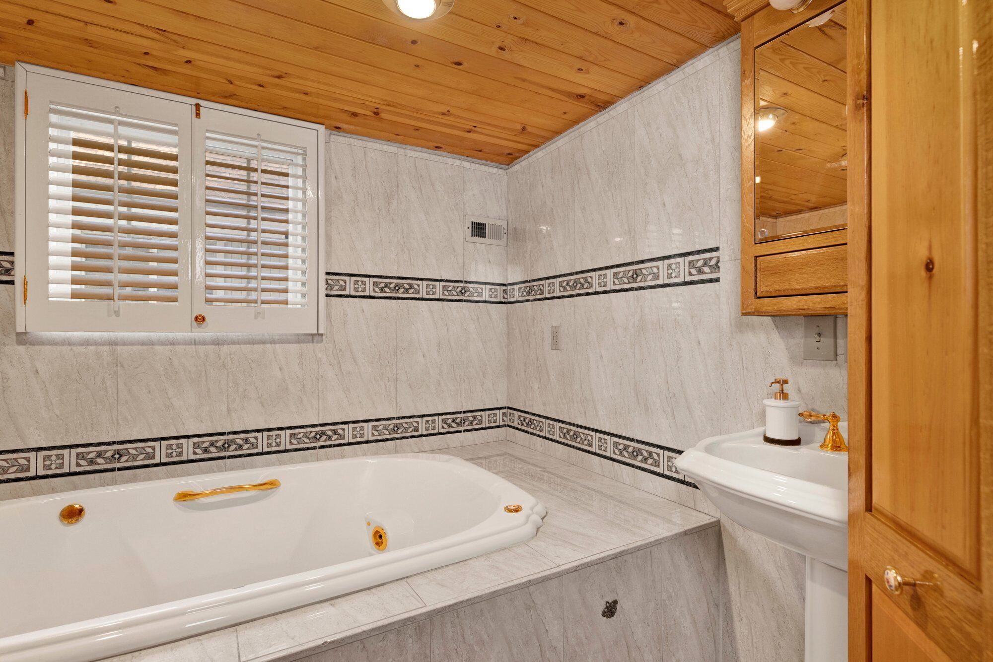 A bathroom with a large bathtub, a sink, wooden ceiling, and a window with blinds.