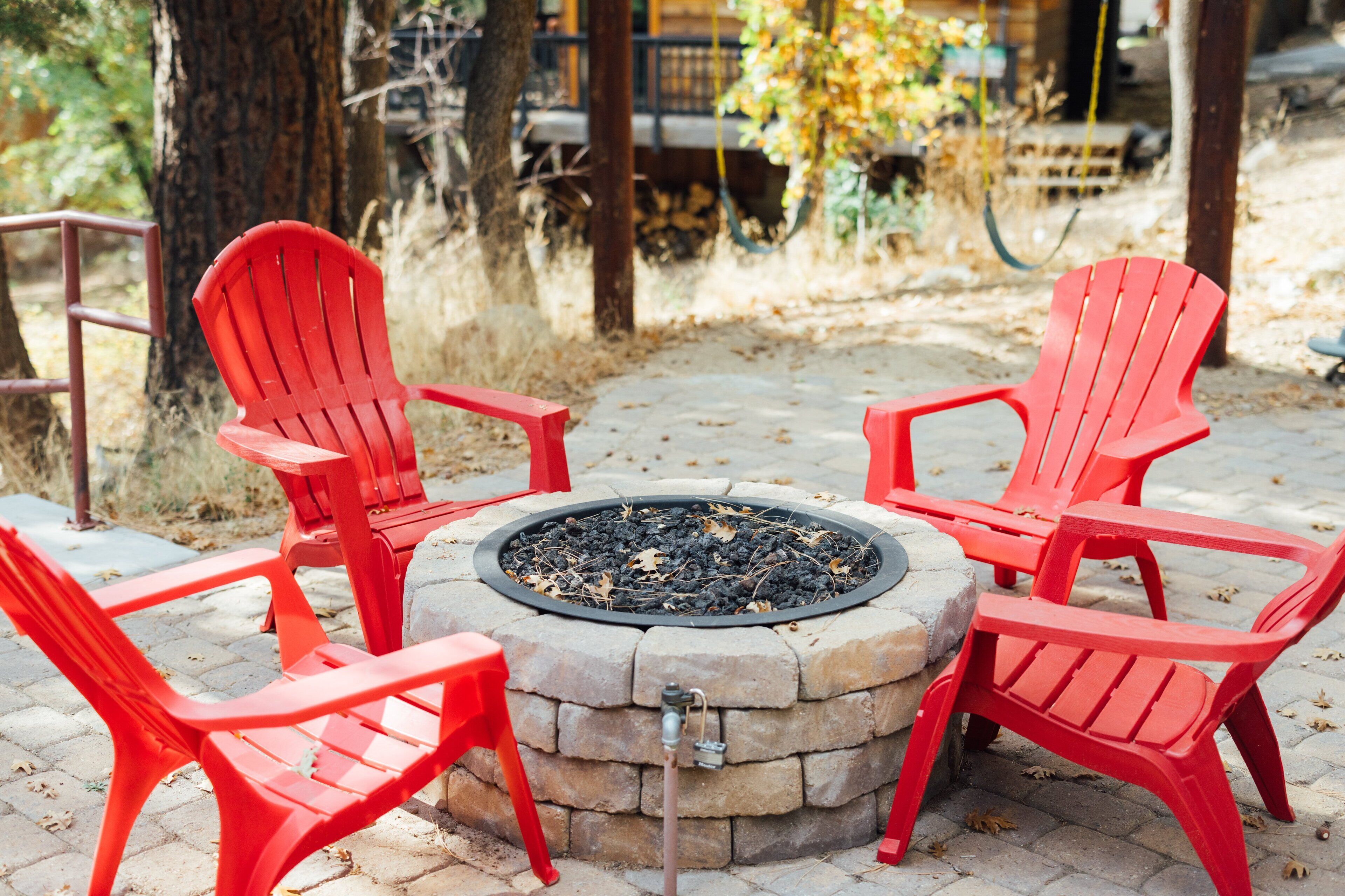 A circular stone fire pit with four red Adirondack chairs around it, set on a paved patio with trees and a wooden deck in the background.