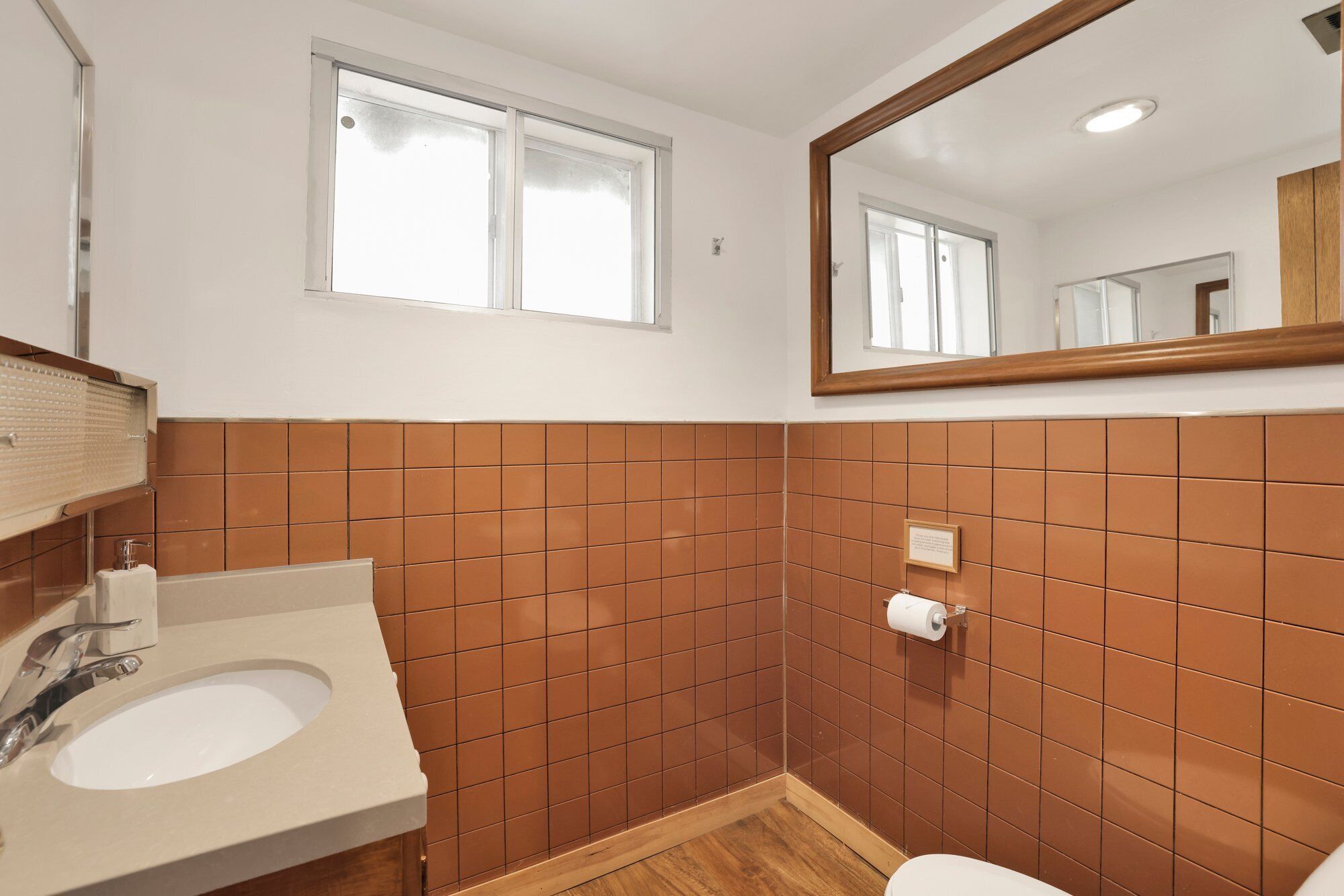Un baño con un lavabo blanco, paredes de azulejos marrones, suelo de madera, una ventana y un espejo.