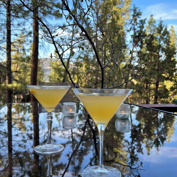 Two martini glasses filled with a yellow beverage on a glass table with a reflection of trees and a clear sky.