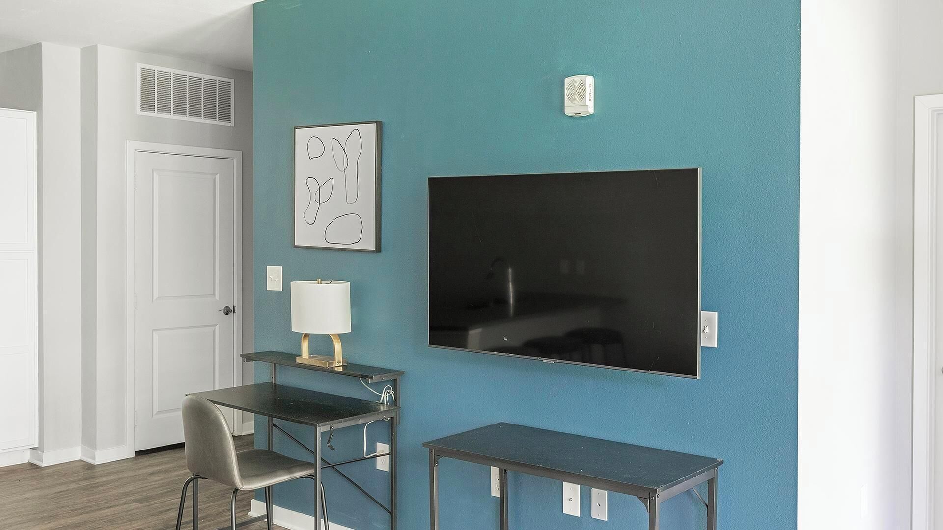 Et lille værelse med teal farvet væg, et stort fladskærms-tv, et lille bord, en stol og en lampe.