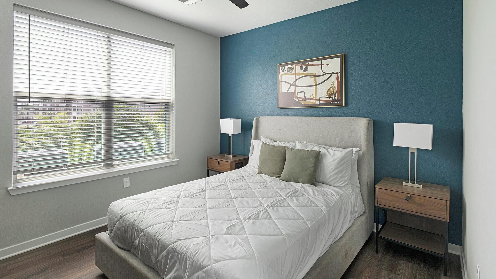A bedroom with a bed, two bedside tables, a window with blinds, and a wall-mounted painting.