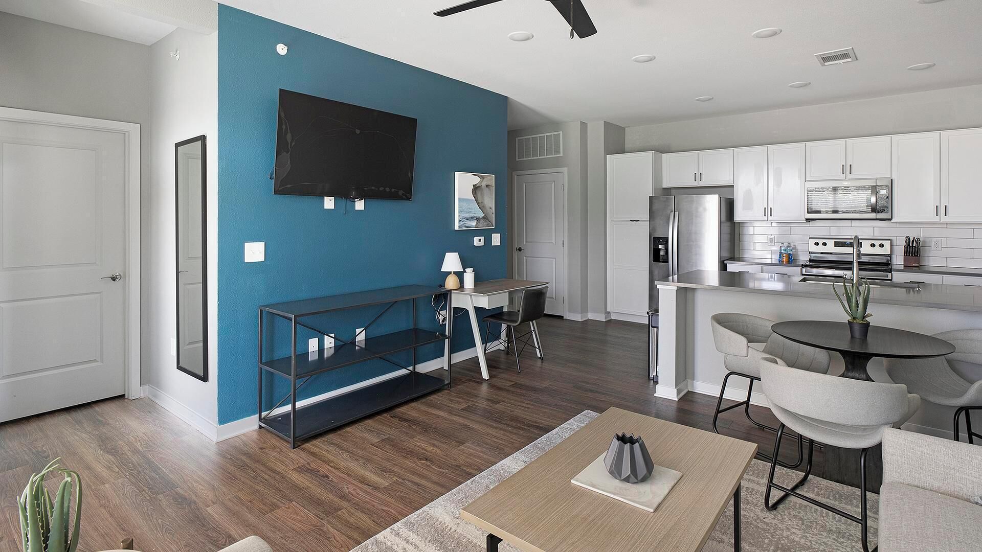 A modern living room with a blue accent wall, a flat-screen TV, a dining area with a round table, and a kitchen with white cabinets and stainless steel appliances.