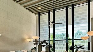 Un área de descanso moderna con un sofá modular, mesas redondas de centro e lámparas de pie. Ventanas amplias ofrecen luz natural y vistas al verdor exterior.